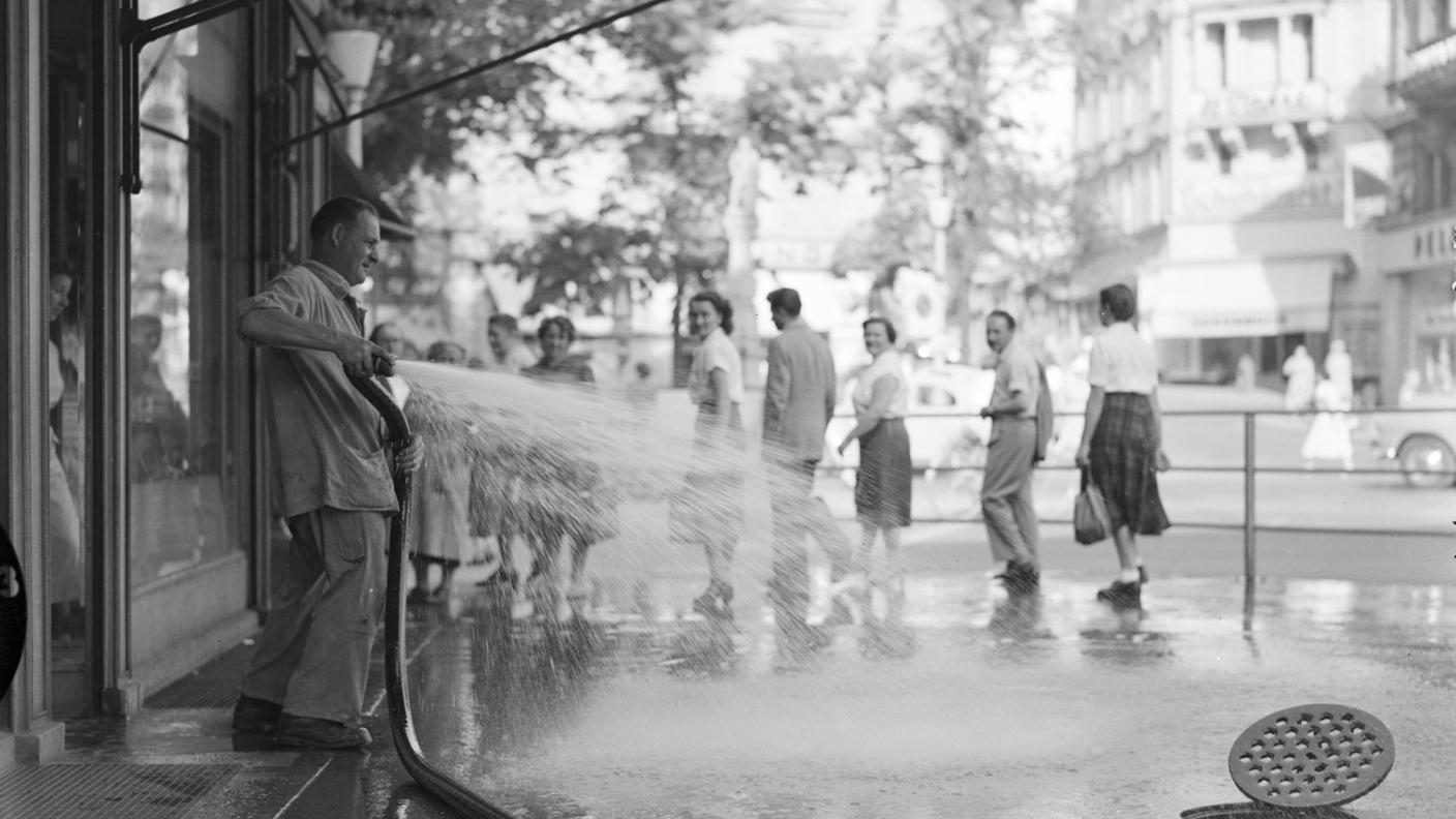 Un uomo usa un tubo flessibile di grosso calibro per spruzzare acqua sull’asfalto davanti al suo negozio, per rinfrescarlo un po’. La foto è stata scattata in una città della Svizzera negli anni ’50, in una calda giornata estiva