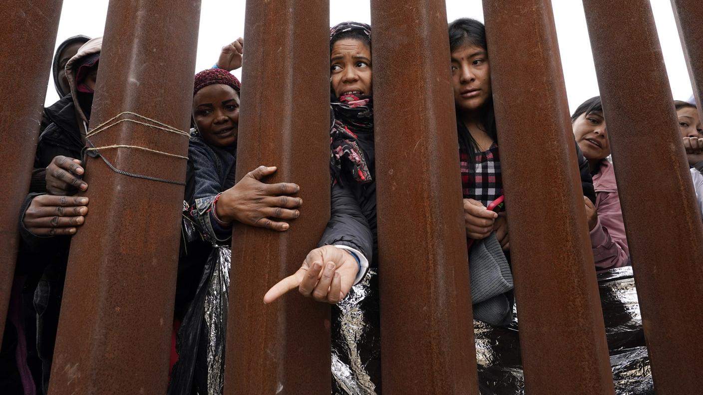 Migranti allungano le braccia attraverso un muro di confine per ricevere vestiti distribuiti dai volontari, 2023, San Diego.