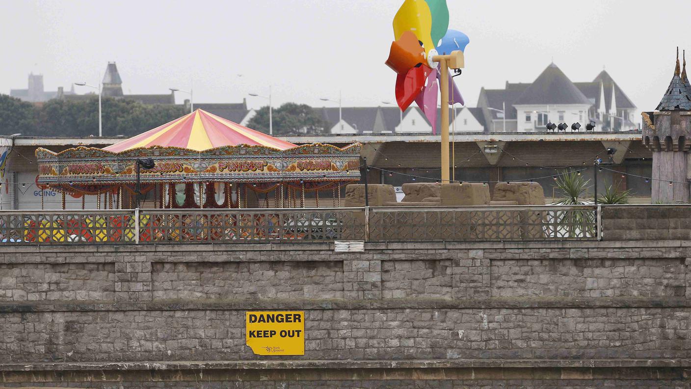 Una giostra si scorge oltre il muro di Dismaland. Il cartello segnala "pericolo"