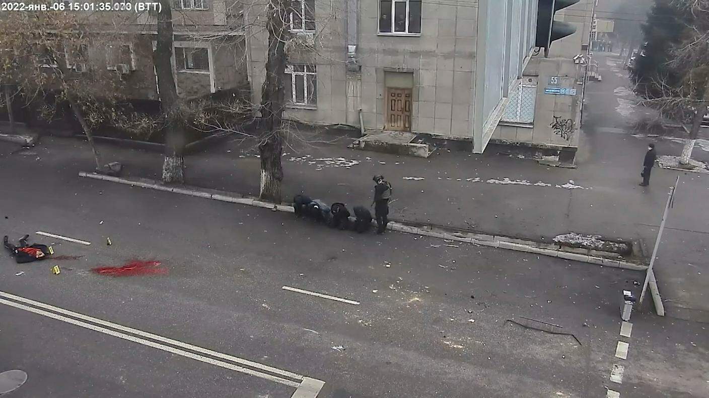 Un cadavere a terra, su una strada di Almaty, mentre un agente sorveglia alcune persone arrestate durante le proteste