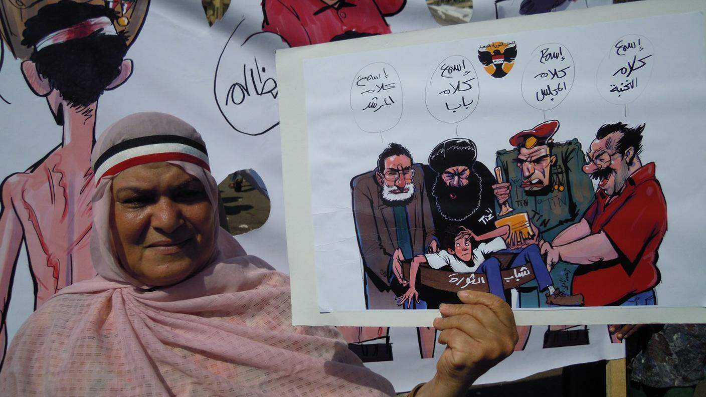 Una donna, in piazza Tahrir nel 2011, mostra con una vignetta la situazione nel Paese: un giovane rivoluzionario strattonato da militari, musulmani e salafiti