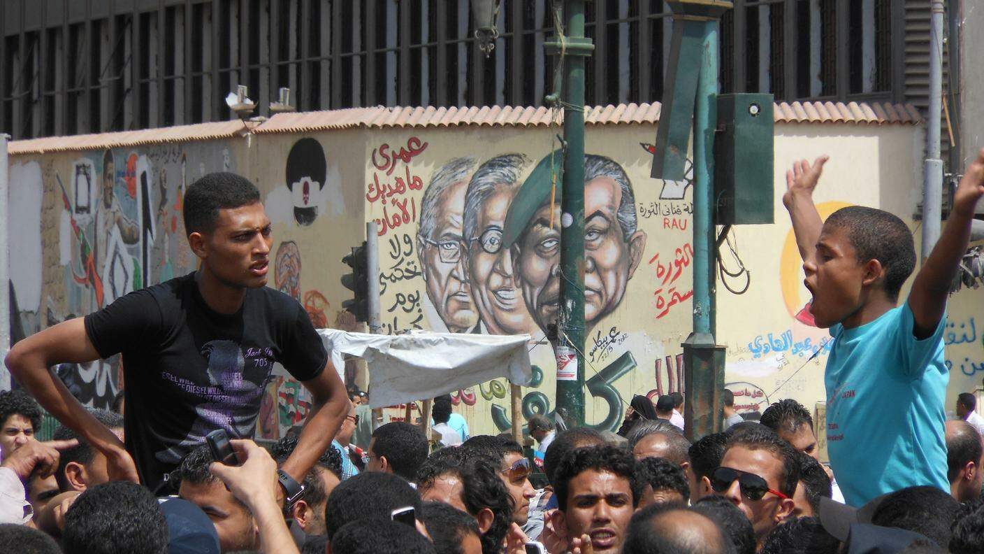 Due ragazzi lanciano i cori in piazza Tahrir, sullo sfondo un murale che raffigura l'era di Mubarak