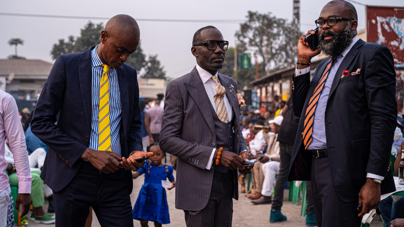 Il sapeur “Derrick” assieme ad altri dandy congolesi conversano nel quartiere Bacongo di Brazzaville. Anche nella Società degli animatori e delle persone eleganti ci sono gruppi e insegnanti seguono i giovani apprendisti. Derrick ha iniziato nel “Clan Piccadilly” detto dei “Diables rouges de la Sape” attorno ai 15 anni.